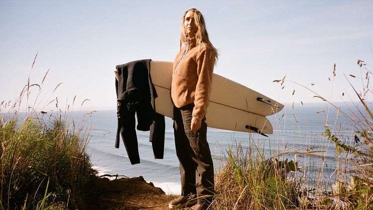 Women surfer in a Billabong fleece holding a surfboard and wetsuit with the ocean behind her.